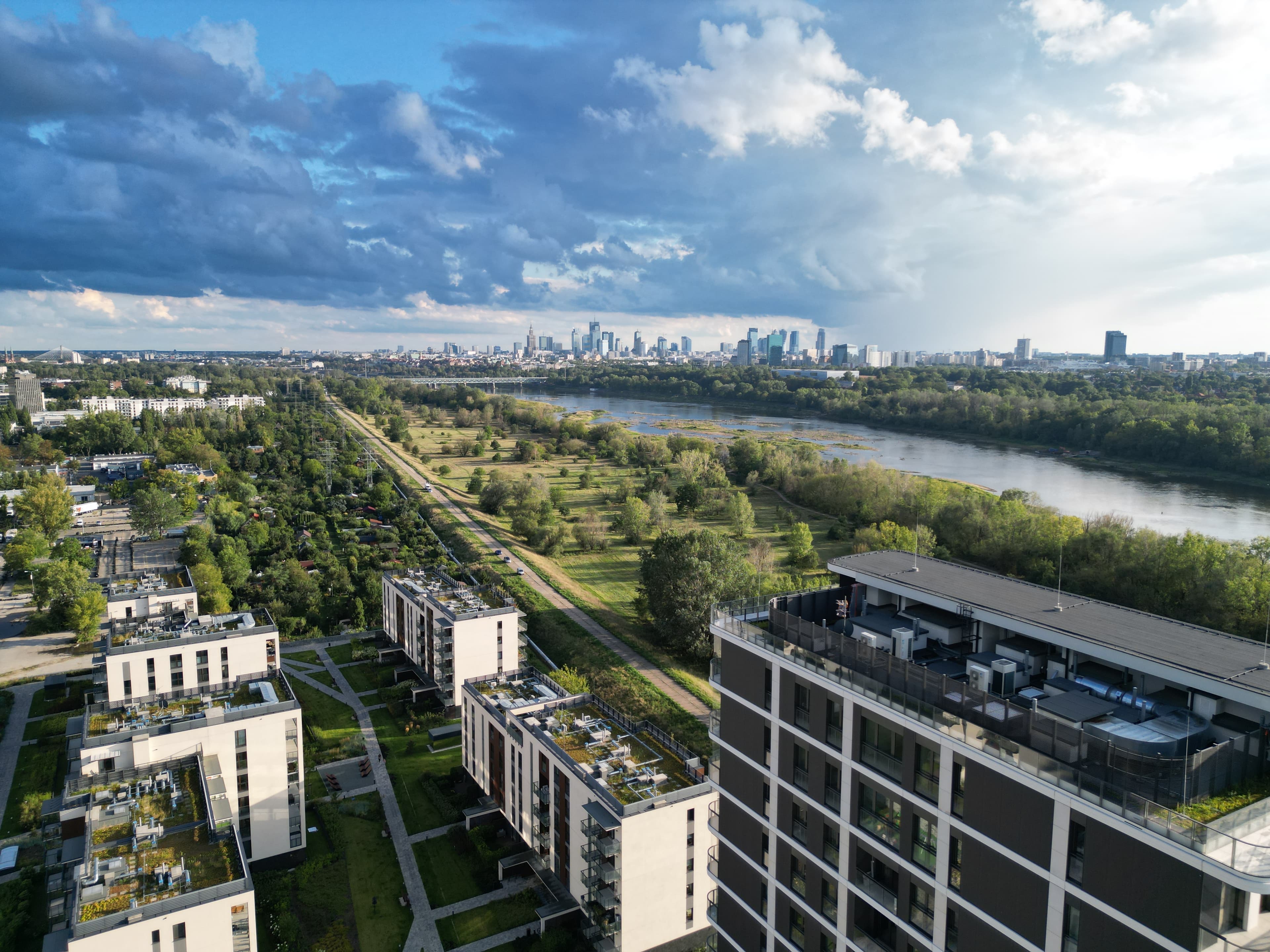 Aerial view of Bulwary Praskie along the Vistula, with the finished blocks, the wild riverbank, and Warsaw’s skyline beyond.