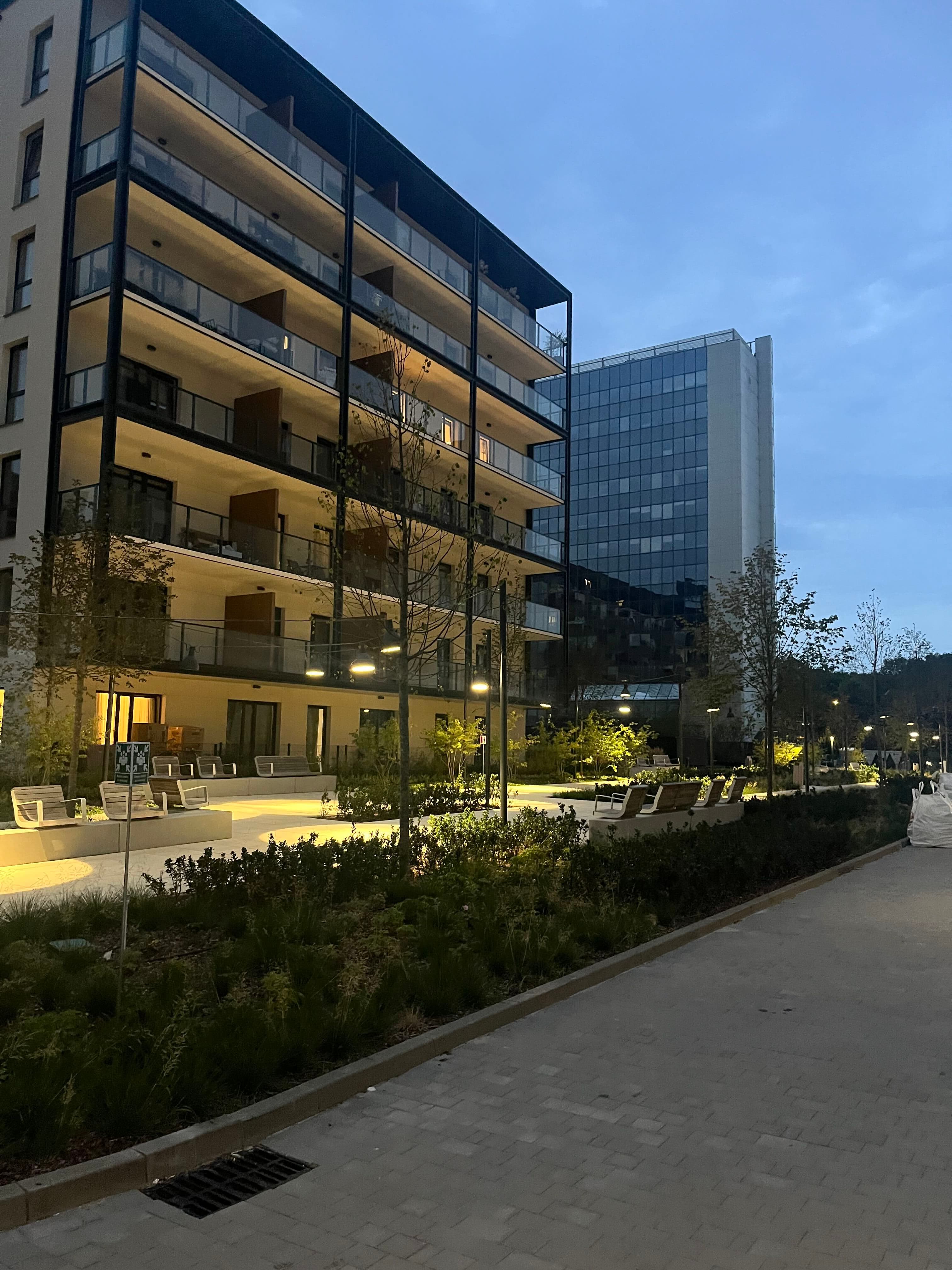 Central social area with curved benches, young trees, and glass balconies at twilight.