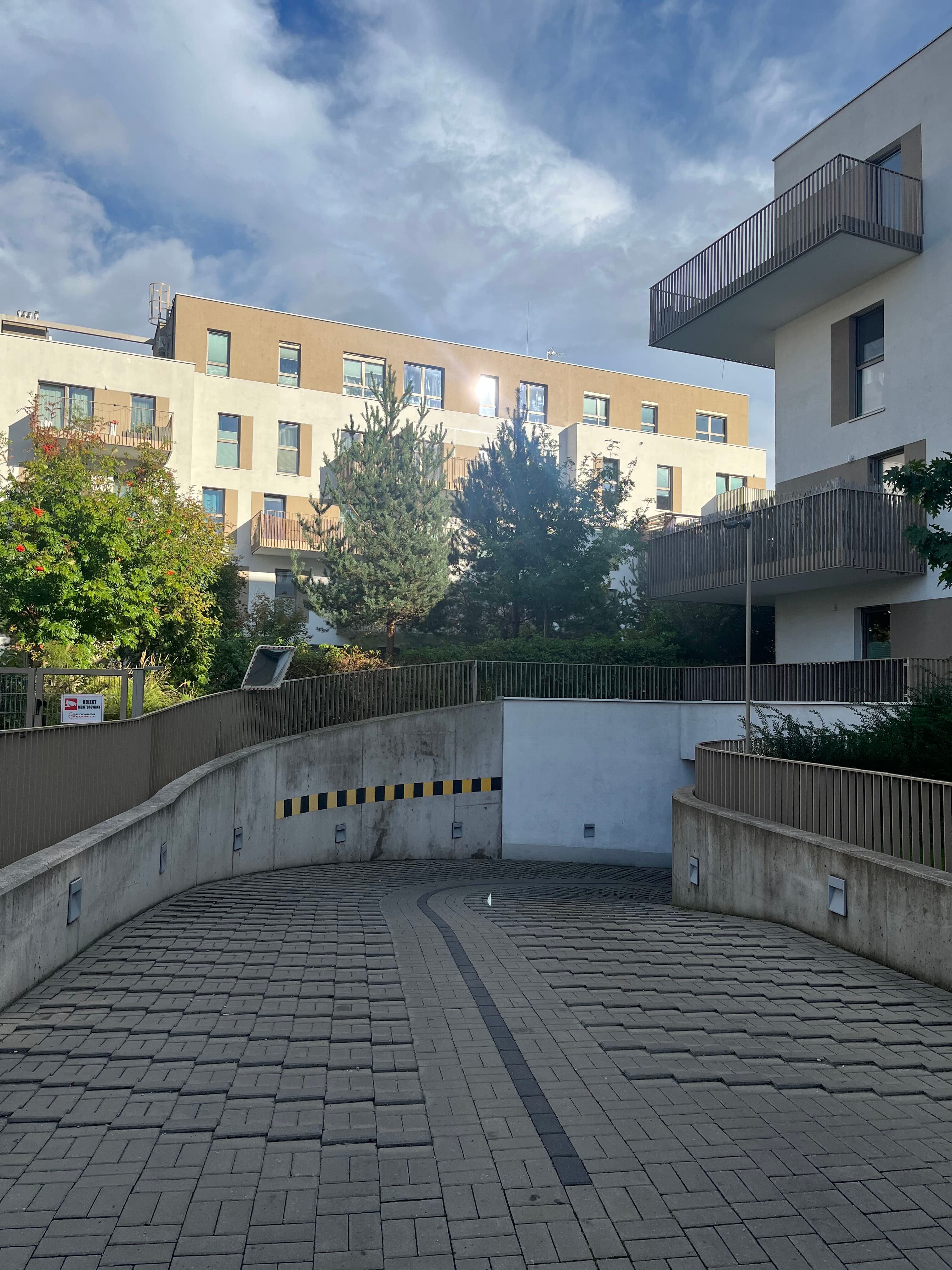 Underground parking ramp with pines and rowan beside white façades and wood-slat balconies.